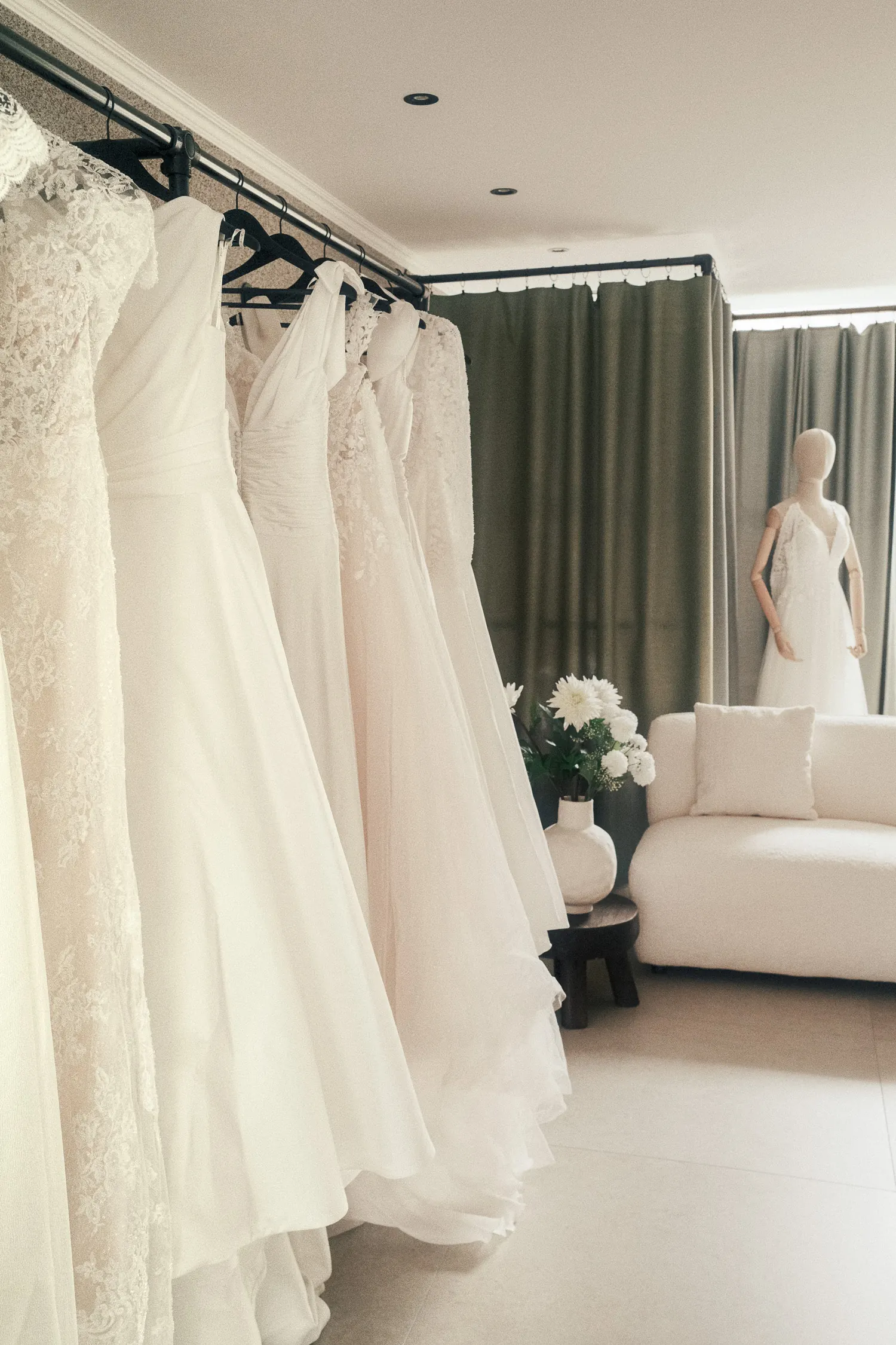 Une sélection de robes de mariée sur un portant, un mannequin habillé au fond de l’atelier